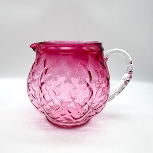 May include: A pink glass pitcher with a clear glass handle. The pitcher has a textured, embossed design and a gradient from a darker pink to a lighter pink.