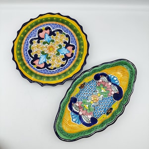 May include: Two colorful, hand-painted ceramic serving dishes. One is round with a scalloped edge, featuring a floral design in yellow, blue, green, and orange. The other is oval, with a similar floral pattern and scalloped edge. Both have a vibrant, decorative style.