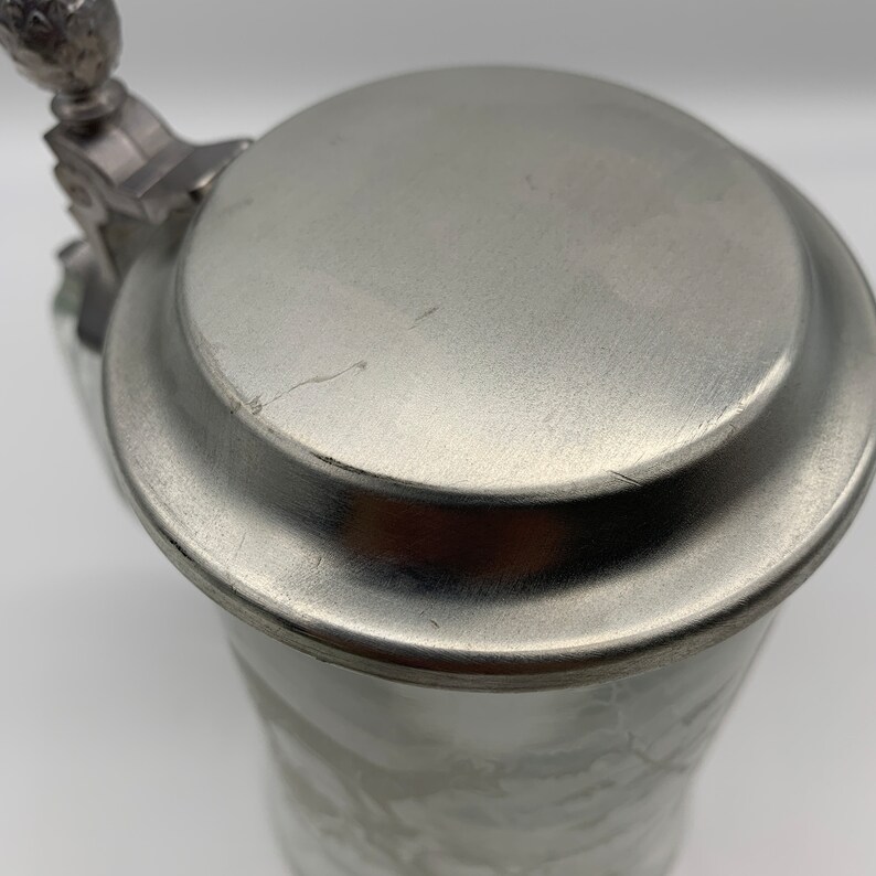 May include: Close-up of the top of a silver beer stein with a lid. The lid has a small, ornate knob on top.