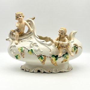 May include: Cream-colored ceramic planter with cherub figures and gold accents. The planter features a boat-like shape with a decorative vine of grapes and leaves. One cherub is seated, and the other is playing a trumpet. A decorative piece for the home.