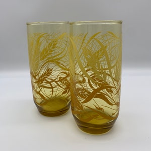 May include: Two tall, yellow-tinted drinking glasses. Each glass features a wheat stalk design in a darker shade of yellow. The glasses are cylindrical with a slightly flared base, perfect for serving beverages.