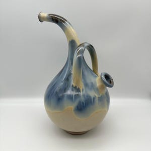 May include: A ceramic vessel with a rounded base and a long, curved spout. The piece features a glossy glaze that transitions from a creamy beige at the bottom to a deep, flowing blue and dark brown pattern across the upper body and handle.