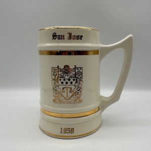 May include: Cream-colored ceramic mug with gold trim and handle. The mug features the text "San Jose" in gold lettering, along with a city emblem and the year "1958". A decorative piece for the home.