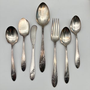May include: An assortment of vintage silverware, including spoons, a fork, and a butter knife, arranged on a light gray surface. The silverware has a floral design on the handles and a shiny, reflective surface.