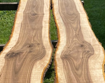 May include: Two long, wide planks of wood with a rich brown and light brown color. The wood has a beautiful grain pattern and is likely walnut. The planks are lying on a wooden surface.