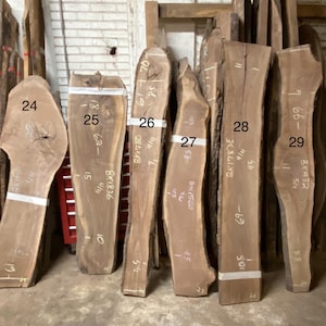 May include: A collection of raw, unfinished wood slabs, varying in shape and size. The slabs are a natural brown color, with visible grain patterns and some knots. Each slab has handwritten measurements and numbers. The slabs are ready for woodworking projects.