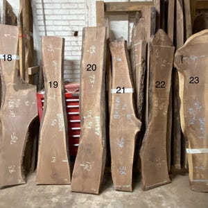 May include: A collection of raw, unfinished wood slabs of varying shapes and sizes. The slabs are a natural brown color, with visible grain patterns. Some slabs have handwritten numbers and measurements in inches.