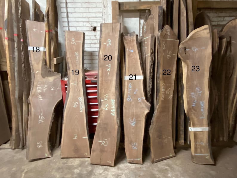 May include: A collection of raw, unfinished wood slabs of varying shapes and sizes. The slabs are a natural brown color, with visible grain patterns. Some slabs have handwritten numbers and measurements in inches.