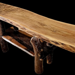 May include: A rustic wooden bench with a natural edge, featuring a light brown tabletop and dark brown, branch-like legs. The bench has a lower shelf and is made of solid wood. The design is reminiscent of a natural, organic aesthetic.