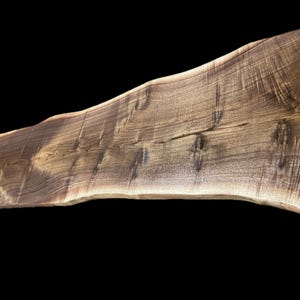 May include: A live edge wooden slab with a natural, irregular edge. The slab is a rich brown color with visible wood grain and knots. The wood appears to be walnut, and the slab is suitable for a table or countertop.