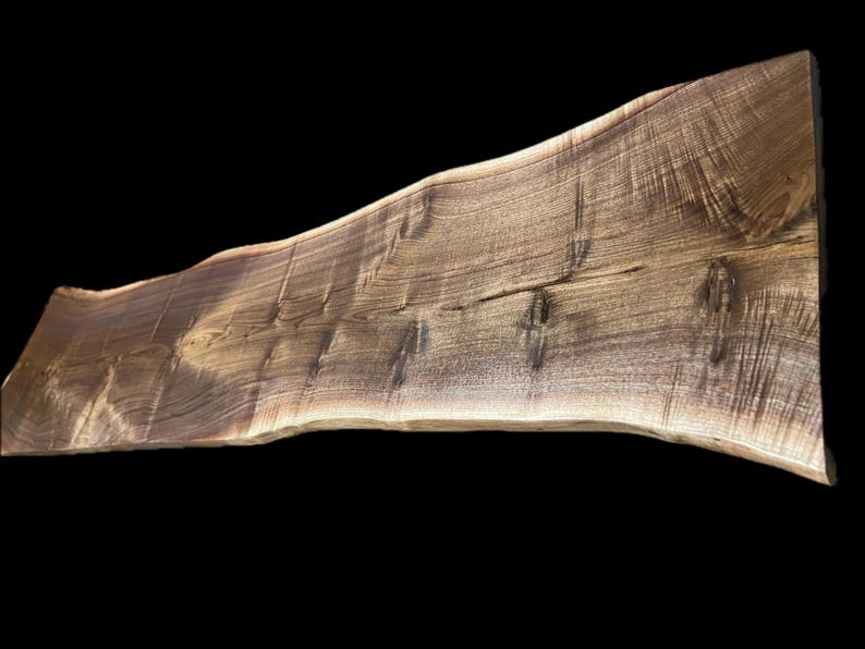 May include: A live edge wooden slab with a natural, irregular edge. The slab is a rich brown color with visible wood grain and knots. The wood appears to be walnut, and the slab is suitable for a table or countertop.