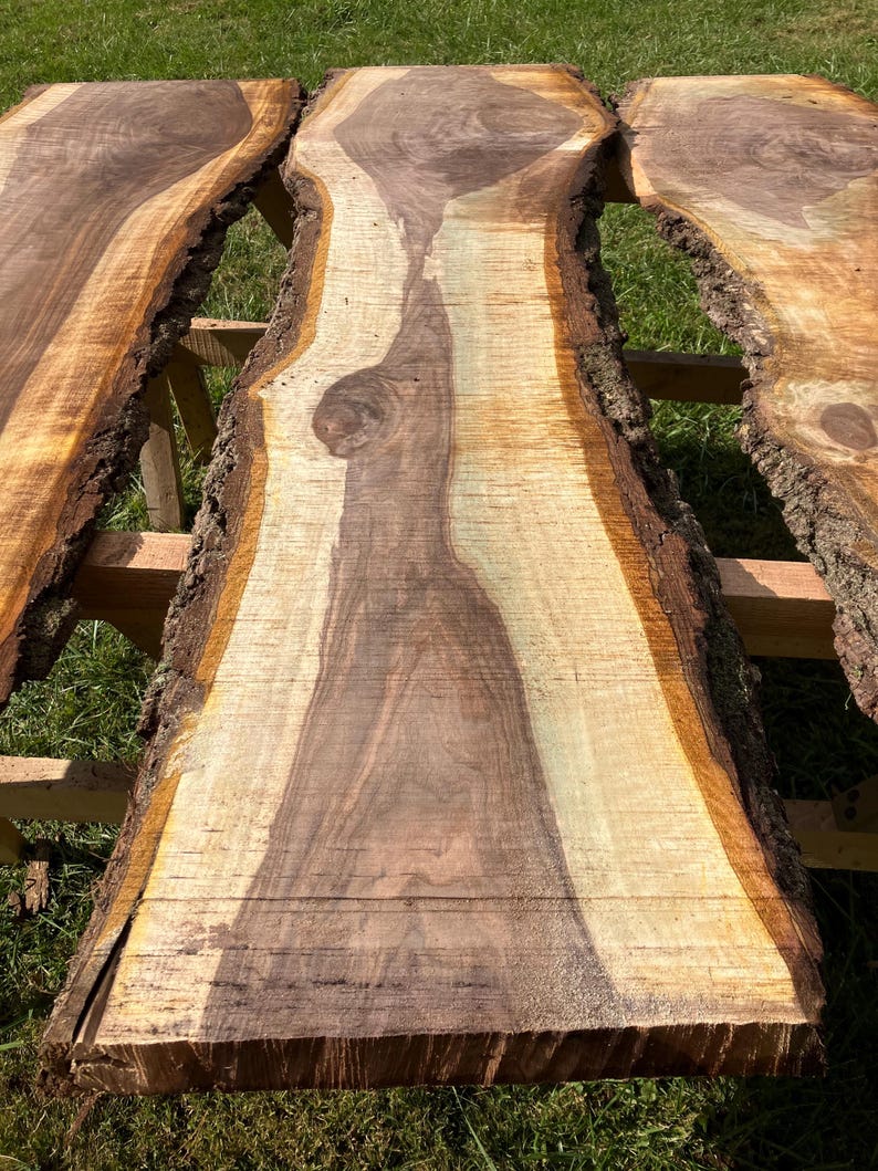 May include: Three unfinished, live-edge wood slabs with natural bark edges. The wood grain features a mix of light and dark brown tones. The slabs are displayed on a wooden support structure, with green grass visible in the background.