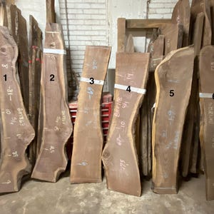 May include: A collection of six unfinished, live edge wood slabs, varying in size and shape. The slabs are a rich brown color, with natural edges and visible grain. Measurements are marked on the slabs in inches.