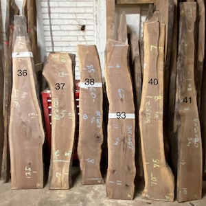 May include: A collection of raw, unfinished wood slabs, varying in size and shape, standing upright. The slabs are numbered and marked with measurements in inches. The wood tones range from light to dark brown, showcasing natural grain patterns.