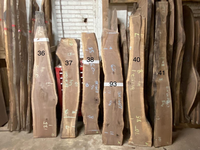 May include: A collection of raw, unfinished wood slabs, varying in size and shape, standing upright. The slabs are numbered and marked with measurements in inches. The wood tones range from light to dark brown, showcasing natural grain patterns.