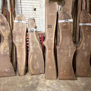 May include: A collection of raw, unfinished wood slabs with natural edges. The slabs vary in shape and size, with visible grain patterns and markings. Some slabs have white tape with numbers and measurements in inches. The wood appears to be a dark brown color.