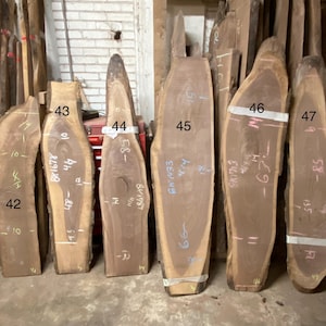 May include: A collection of unfinished wood slabs, varying in shape and size. The slabs are numbered and marked with measurements in inches. The wood has a natural, light brown color with visible grain patterns. Some slabs have a live edge.