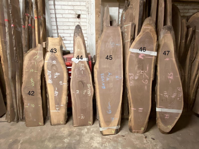 May include: A collection of unfinished wood slabs, varying in shape and size. The slabs are numbered and marked with measurements in inches. The wood has a natural, light brown color with visible grain patterns. Some slabs have a live edge.