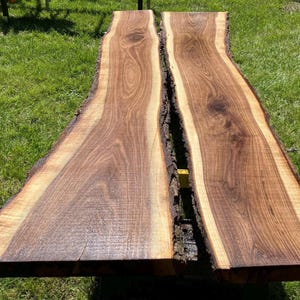 May include: Two live edge wood slabs with a natural edge. The slabs are a rich brown color with lighter tan edges. The wood grain is visible. The slabs are likely for a table or other furniture.