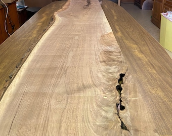 May include: A close-up of a large, light brown wooden table top with a natural edge. The wood has a unique grain pattern and a crack running down the center.