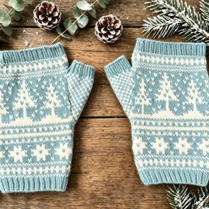 May include: A pair of fingerless gloves in a light blue and white knit pattern featuring a repeating design of evergreen trees and snowflakes. The gloves have ribbed cuffs at the top and bottom edges and a thumb opening on the side of each piece.