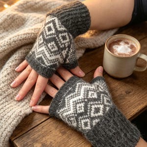 Könnte beinhalten: Ein Paar fingerlose Handschuhe mit einem geometrischen Muster in Dunkelanthrazit und Weiß. Die Handschuhe haben ein geripptes Bündchen am Handgelenk und eine gerippte Öffnung für die Finger, gefertigt aus weichem, flauschigem Strick mit Zickzackmuster.