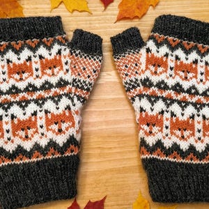 May include: A pair of fingerless gloves featuring a knitted pattern of orange, white, and black fox faces. The gloves have dark charcoal gray ribbed cuffs at the top and bottom, with a repeating geometric design separating the rows of fox motifs.