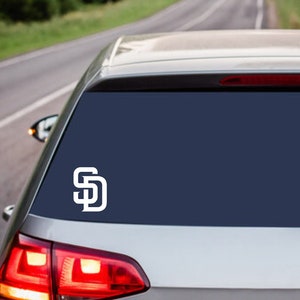May include: White vinyl decal of the San Diego Padres logo on a car window. The logo features the intertwined letters "SD" in a clean, modern design. The car is silver, with a dark blue window.