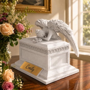 May include: A white stone memorial urn featuring a sculpted angel with detailed wings resting its head on the lid. The rectangular base has a decorative border and a gold-colored metal plaque engraved with the name Emily in black script.