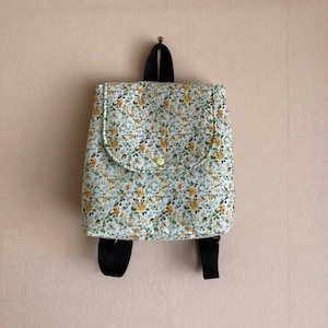 May include: A small backpack featuring a floral design of yellow flowers and green leaves on a white backdrop. It has a flap closure with a yellow button, black straps, and a black handle. The bag is suspended on a wall.