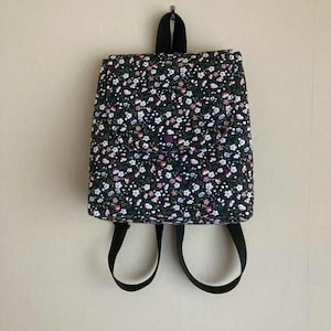 May include: A small backpack with a black floral pattern, featuring tiny white, pink, and purple flowers. It has a flap closure and black adjustable straps. The bag is hanging on a wall.