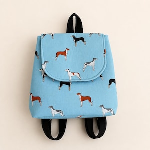 May include: A light blue backpack featuring a repeating pattern of various sighthound dogs in black, white, and brown. The bag has a top flap closure, a black top handle, and two black adjustable shoulder straps attached to the bottom.