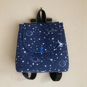 May include: A small, navy blue backpack featuring a celestial design of white stars, planets, and shooting stars. It has a flap secured by a blue button, black straps, and metal buckles.