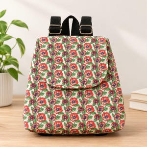 May include: A small backpack featuring a cream-coloured fabric base with a repeating pattern of red and purple flowers on green stems. The bag includes a top flap closure and black adjustable nylon straps with silver-toned metal hardware buckles.