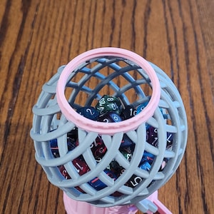 May include: A gray and pink dice tower with a clear plastic top. The tower is filled with colorful dice.