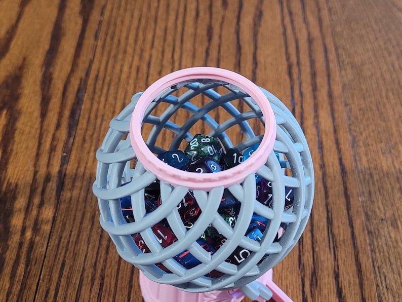 May include: A gray and pink dice tower with a clear plastic top. The tower is filled with colorful dice.
