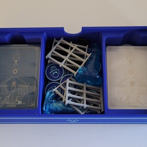 May include: A blue plastic game organiser with three compartments. The compartments contain cards with the word "Hades" printed on them, blue plastic game pieces, and grey plastic game pieces.