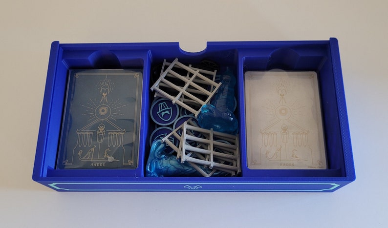 May include: A blue plastic game organiser with three compartments. The compartments contain cards with the word "Hades" printed on them, blue plastic game pieces, and grey plastic game pieces.