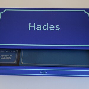 May include: A blue box with a light blue border and the word "Hades" printed on the top. The box is open and the inside is lined with a dark blue fabric. A small card with the text "Hades Objective: Start your turn with three Titans at Mount Olympus" is visible inside the box.