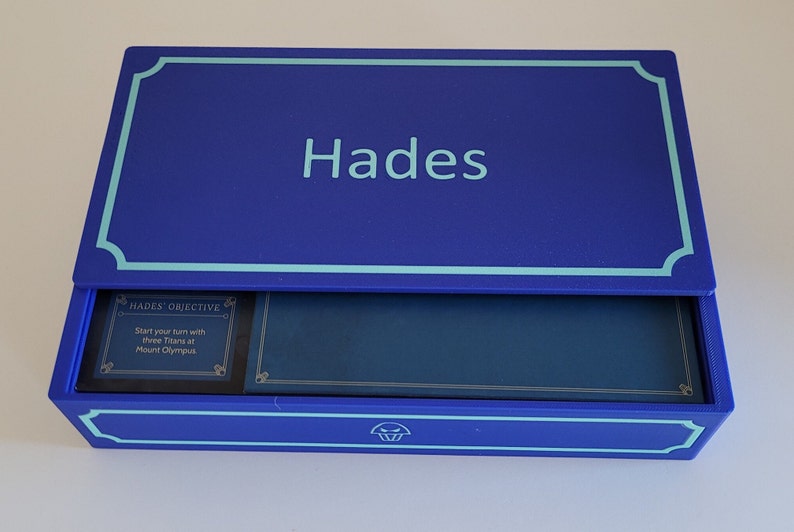 May include: A blue box with a light blue border and the word "Hades" printed on the top. The box is open and the inside is lined with a dark blue fabric. A small card with the text "Hades Objective: Start your turn with three Titans at Mount Olympus" is visible inside the box.