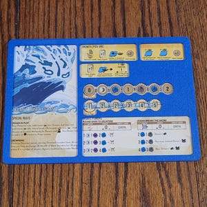 May include: A blue game board with various game elements. The board features text and graphics related to a coastal theme, including the words "Ocean in Play" and "Ocean Breaks the Shore". The board is on a wooden surface.
