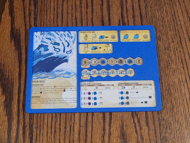 May include: A blue game board with various game elements. The board features text and graphics related to a coastal theme, including the words "Ocean in Play" and "Ocean Breaks the Shore". The board is on a wooden surface.