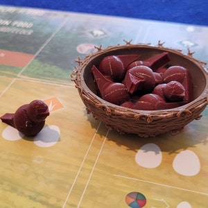 May include: A brown bird figurine and a brown woven nest filled with red plastic eggs. The nest and eggs are part of a board game.