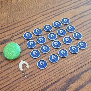 May include: A collection of themed game pieces, including a green oval stone with a spiral design, a bone-colored hook, and several blue and white circular tokens with a hook symbol. The items are arranged on a wooden surface.