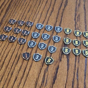May include: A collection of small shield-shaped tokens arranged in three groups. The copper-colored tokens are marked with the number 1, the silver-colored tokens are marked with the number 3, and the gold-colored tokens are marked with the number 5.