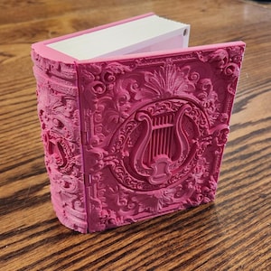 May include: A magenta-coloured decorative book with detailed, embossed patterns. The cover displays a lyre within a circular frame, surrounded by floral and scrollwork designs. The pages are white, and the book is open, revealing the interior.
