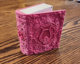 May include: A pink, decorative book with ornate, embossed detailing. The cover features a central lyre design within a circular frame, surrounded by floral and scrollwork patterns. The pages are white, and the book is open, revealing the interior.
