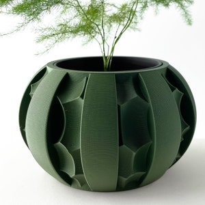 May include: A green ceramic planter with a geometric pattern. The planter is round and has a wide opening. A small green plant is growing inside the planter.