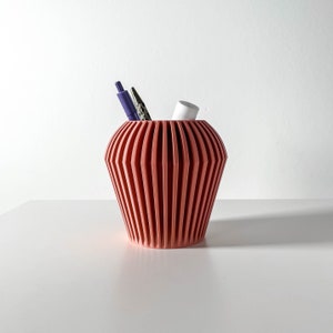 Può includere: Un organizzatore da scrivania in ceramica color corallo con un design geometrico. L'organizzatore ha una base larga e arrotondata e una parte superiore stretta. Due penne e un pennarello bianco sono all'interno dell'organizzatore.