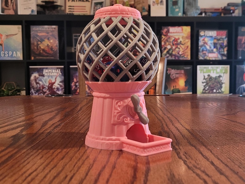 May include: A pink and gray tabletop dice tower with a crank handle. The tower is shaped like a vintage gumball machine. The dice tower is filled with various colored dice.